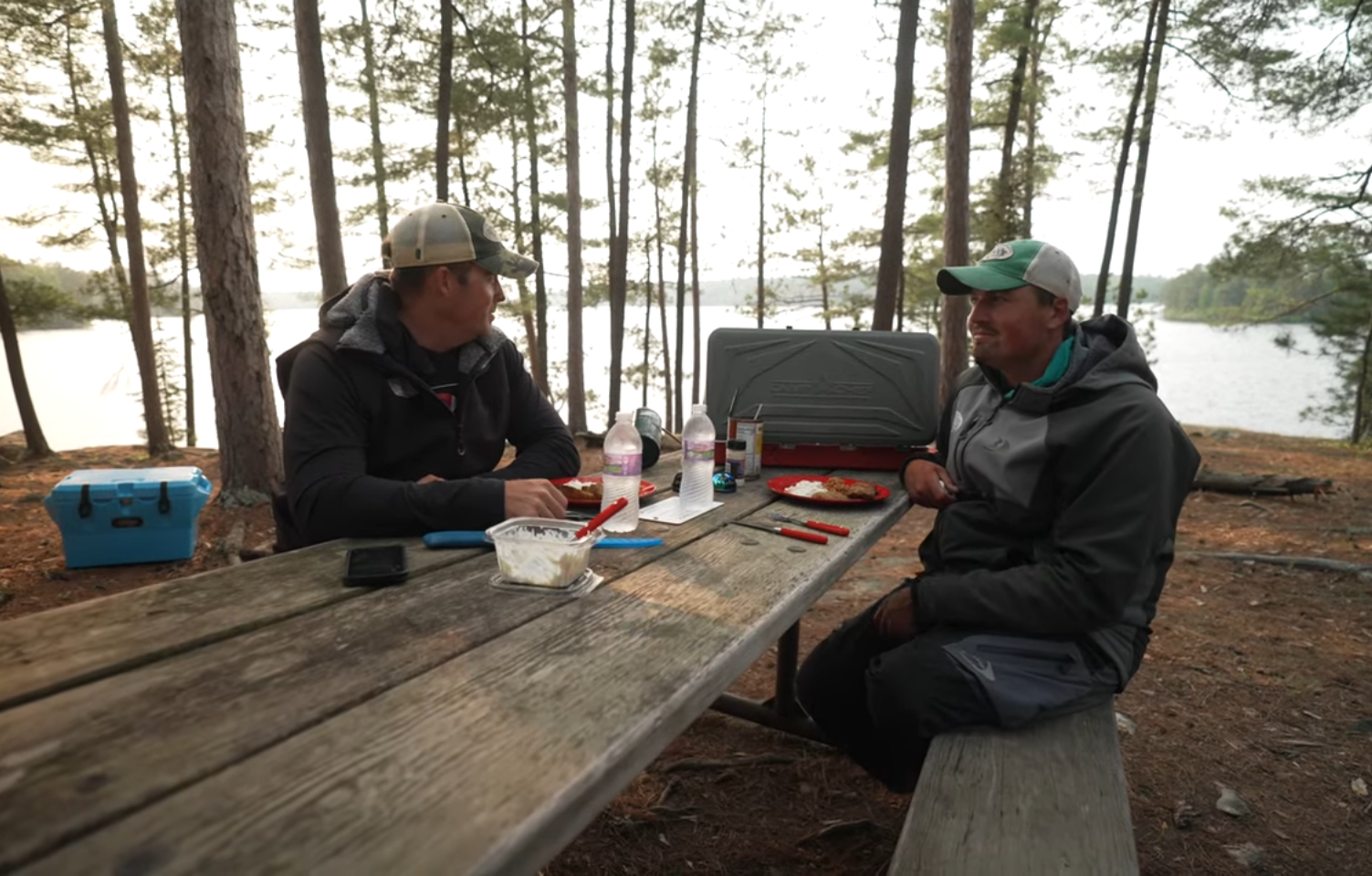 Two people in outdoor jackets sit at a wooden picnic table with food and drinks, surrounded by tall trees near a lake—a fishing guide rests nearby. Cooking supplies and a portable stove are on the table, with a blue cooler close at hand.
